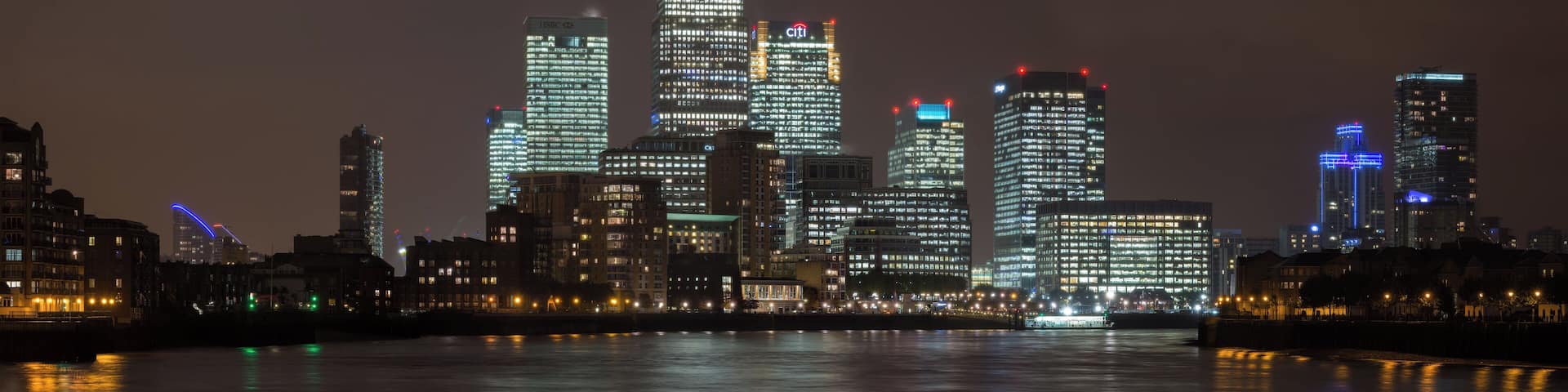 A nighttime city skyline with illuminated buildings reflecting on the water.