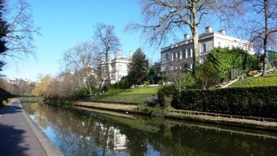 A calm river with a pathway alongside, a large building with a garden, and leafless trees.