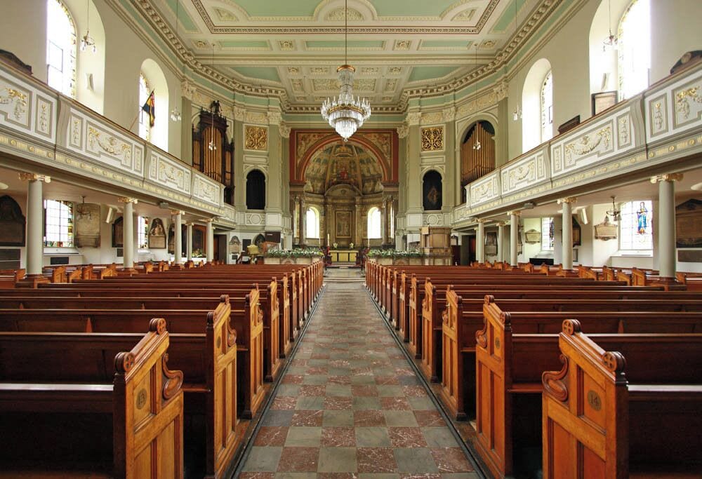 Interior de igreja com fileiras de bancos de madeira, um corredor central e um candelabro.