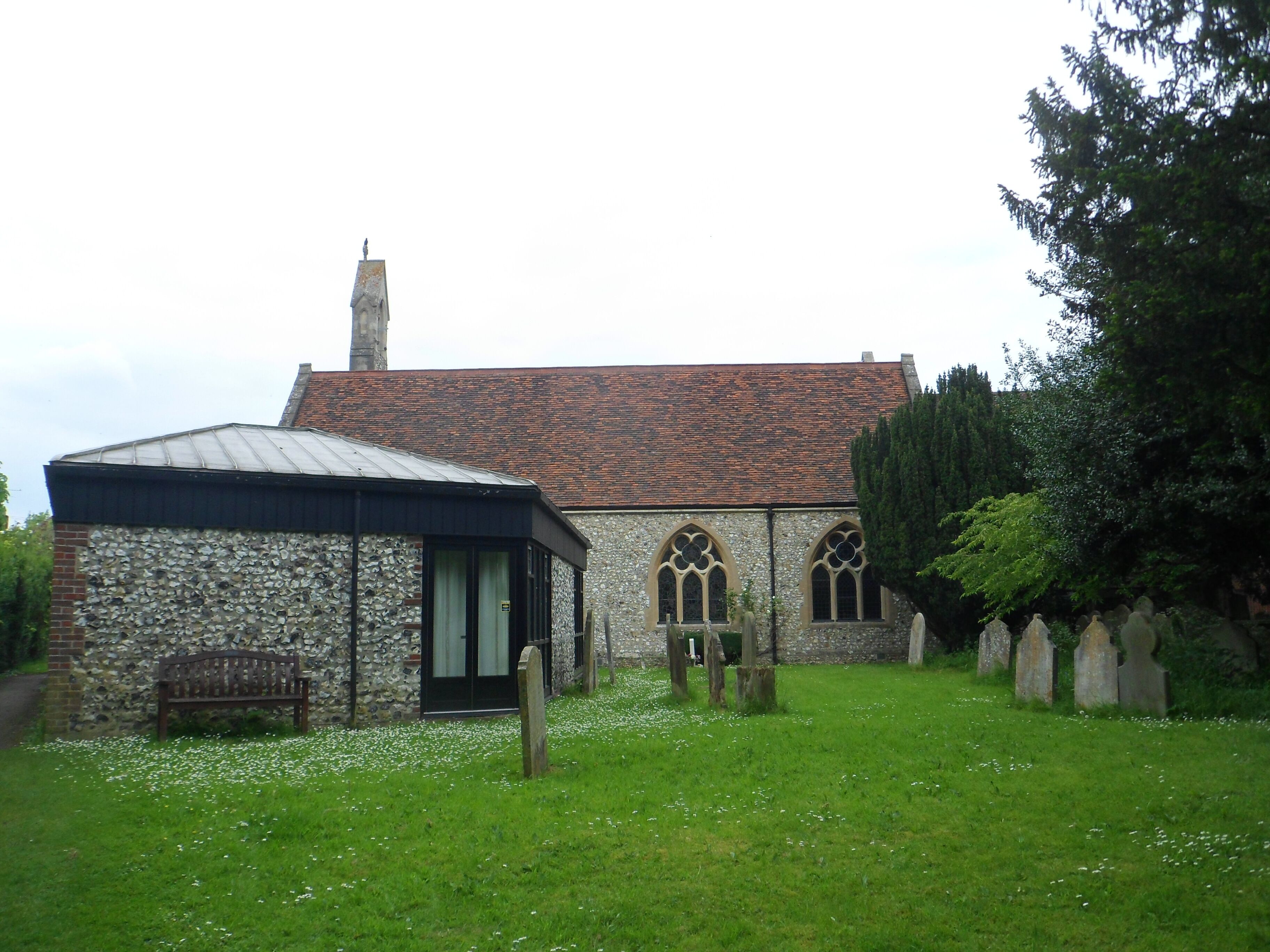 A stone church with a tiled roof, a wooden bench, and a grassy area with white flowers.