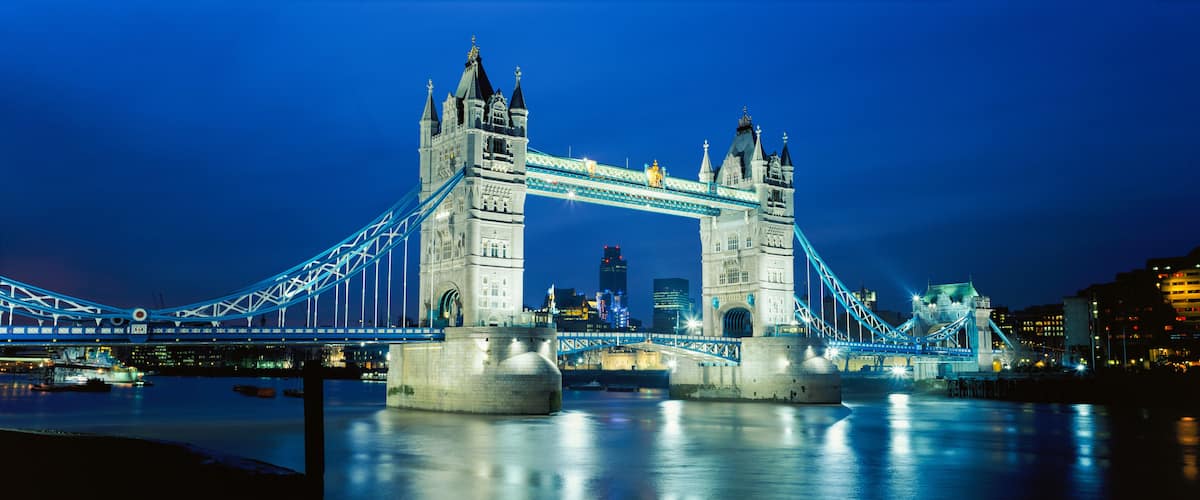 The Tower Bridge at night with illuminated blue lights, a river in the foreground, and a city skyline in the background.