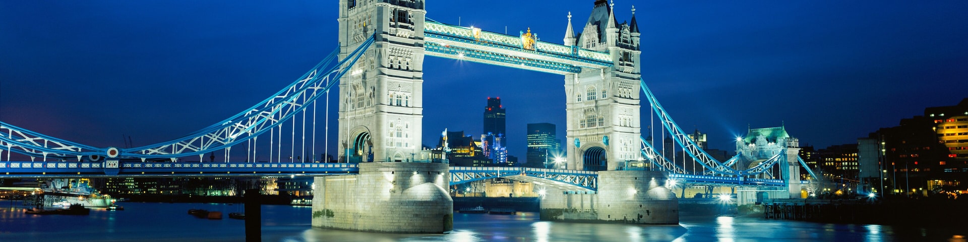 The Tower Bridge at night with illuminated blue lights, a river in the foreground, and a city skyline in the background.