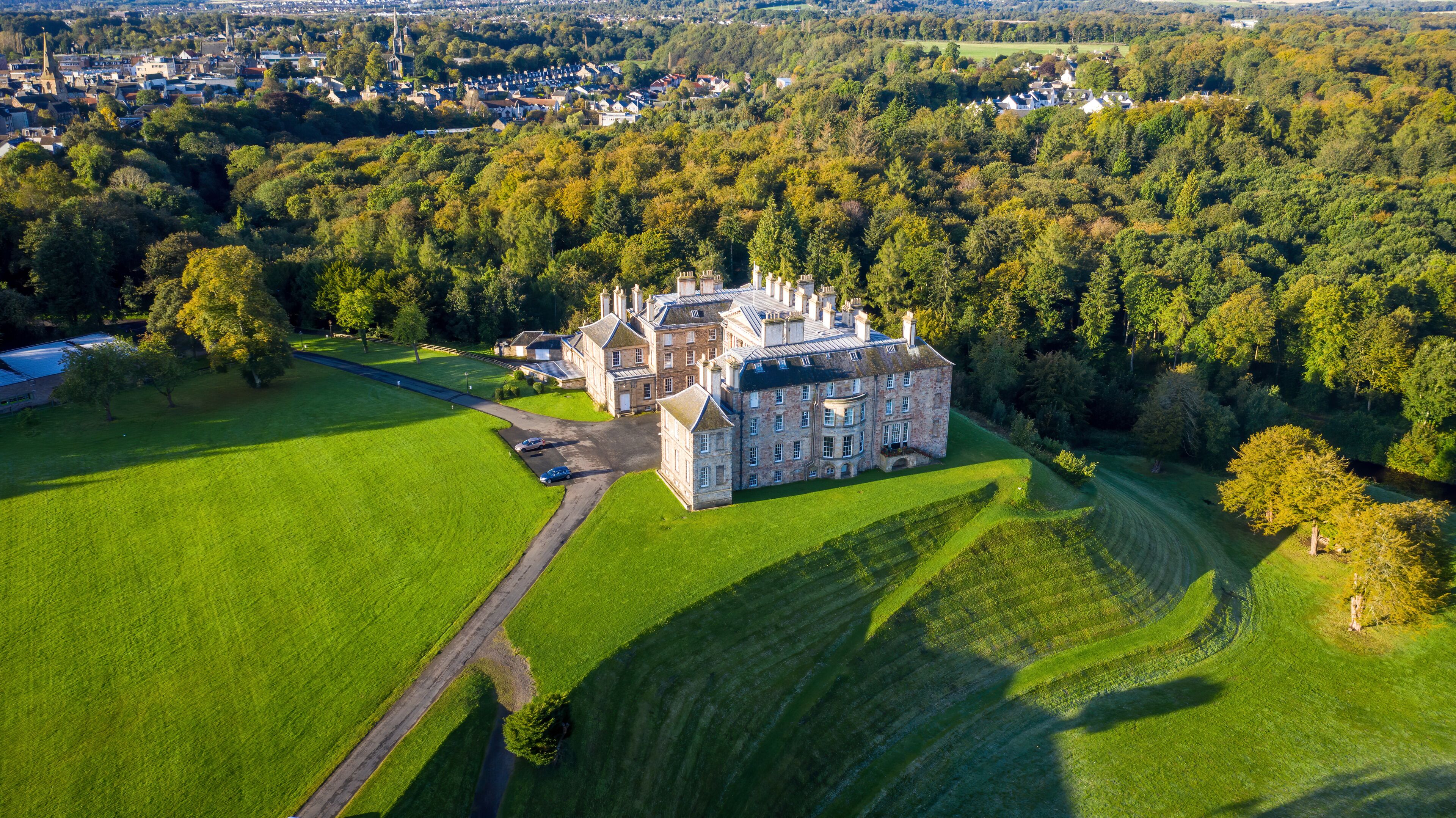 An aerial view of a large estate with a prominent castle-like structure surrounded by lush green fields and dense forests.