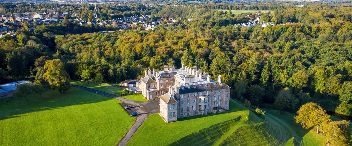 An aerial view of a large estate with a prominent castle-like structure surrounded by lush green fields and dense forests.