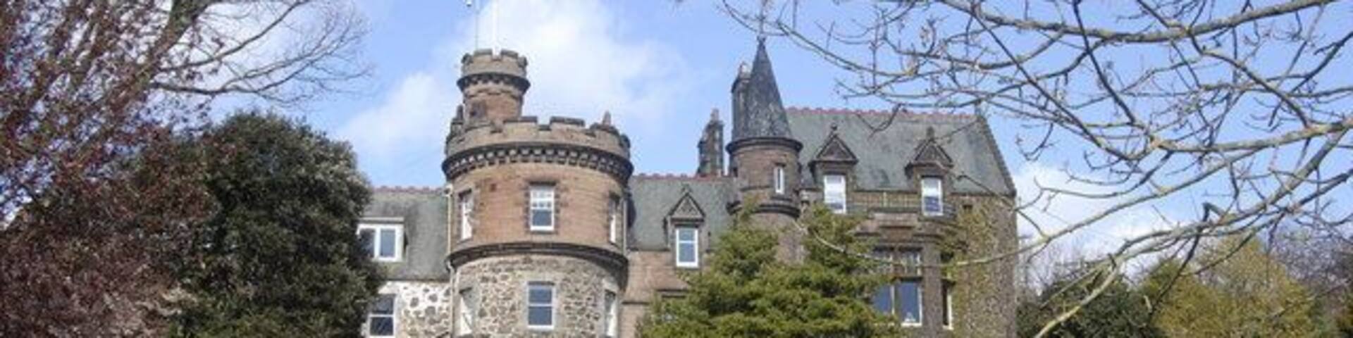 A large, historic building with a turret and multiple chimneys, surrounded by a well-maintained garden and trees.