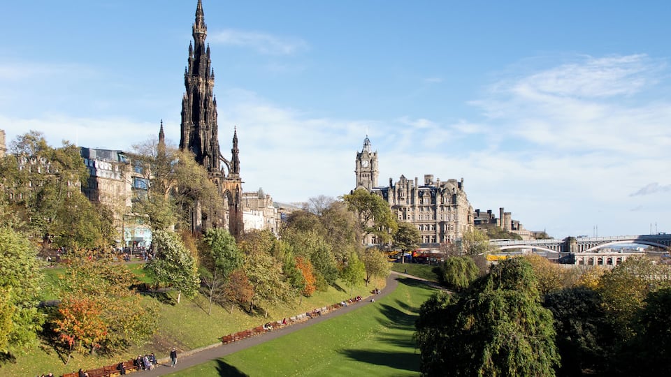 A historic cityscape with a prominent cathedral and a clock tower, surrounded by lush green parks and modern buildings.