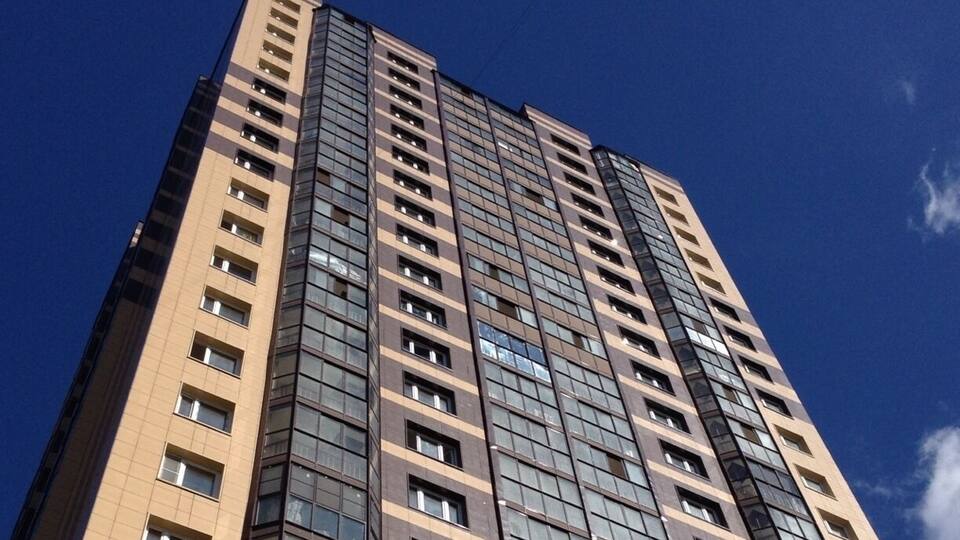 A tall, modern high-rise building with a beige and brown facade, numerous windows, and a clear blue sky in the background.