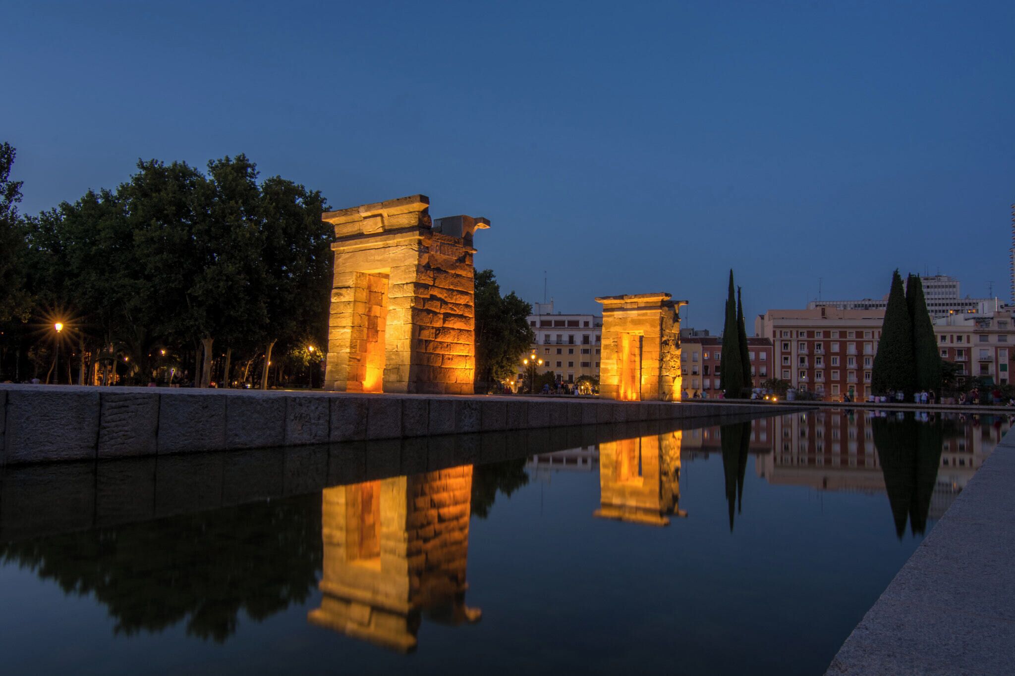 A nighttime view of ancient stone structures by a calm water body, reflecting the buildings and trees.