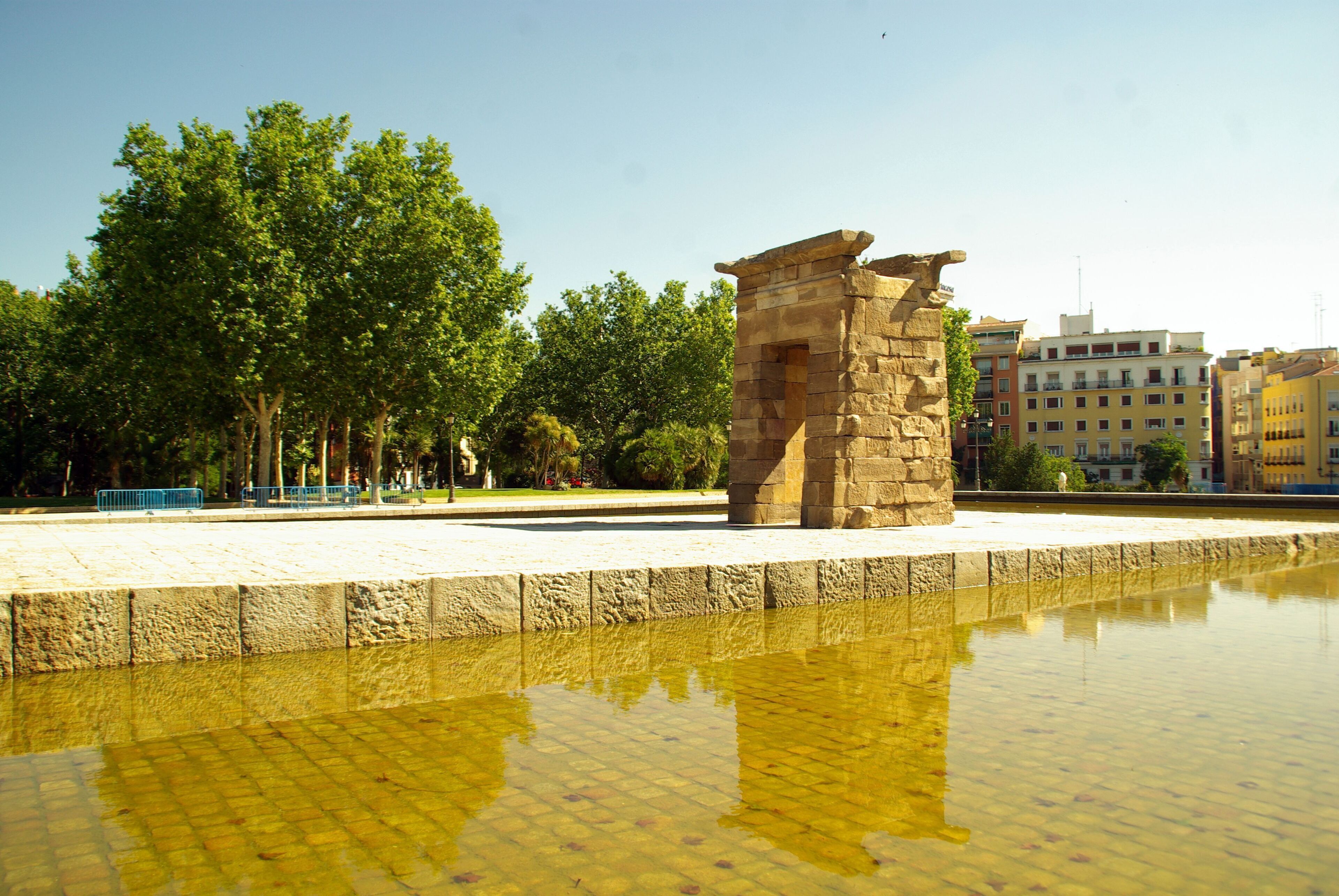 A stone structure in a park with a reflective water surface, surrounded by trees and buildings.