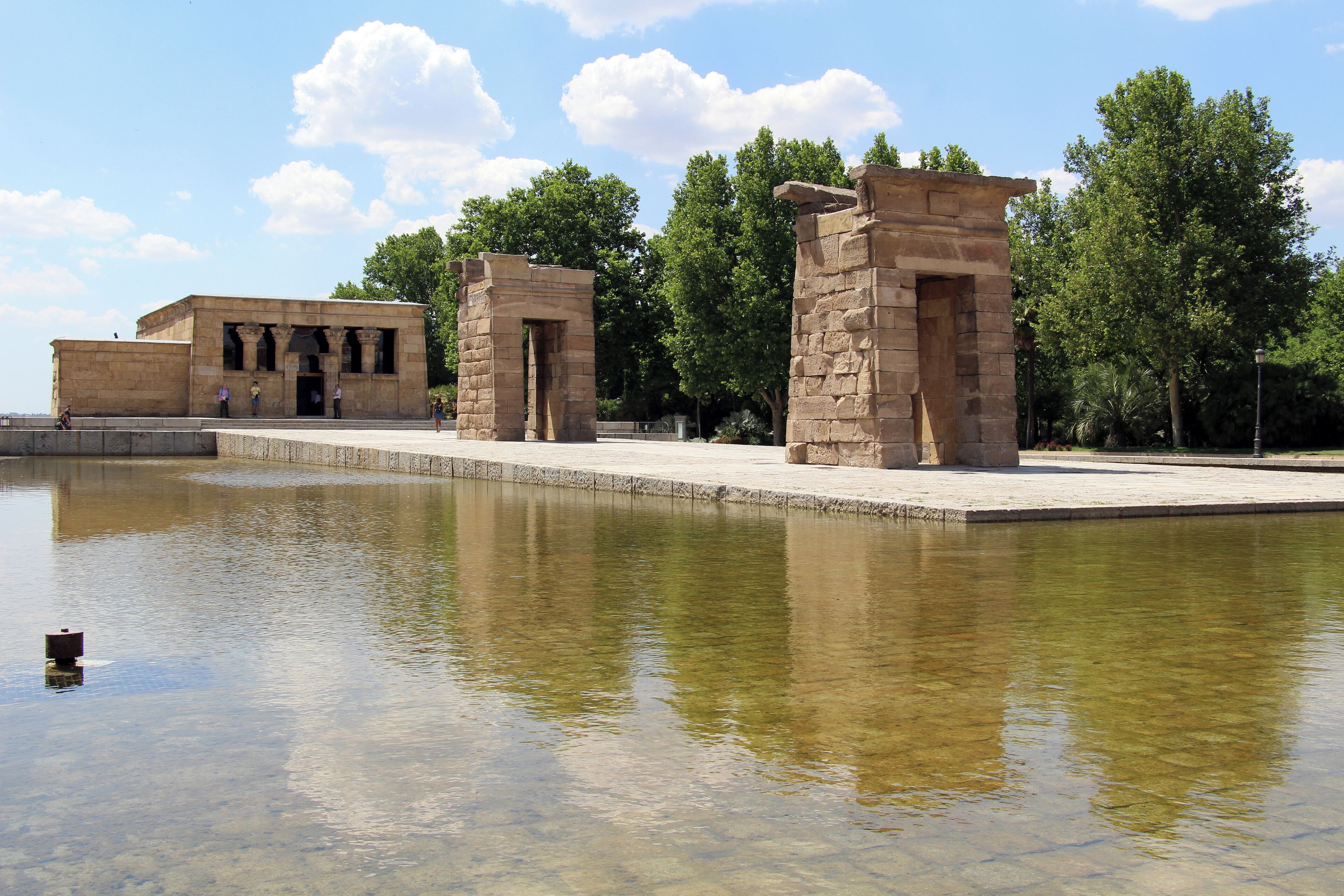 Ancient stone structures by a reflective water body, surrounded by trees.