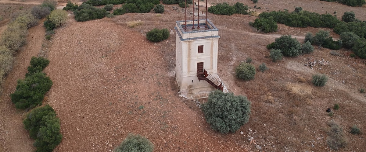 An isolated tower amidst a dry, hilly landscape with sparse vegetation and distant industrial structures.