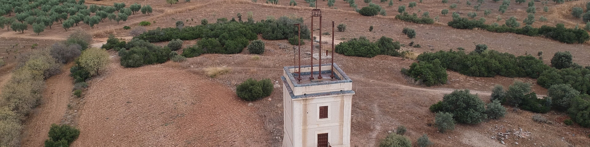 An isolated tower amidst a dry, hilly landscape with sparse vegetation and distant industrial structures.