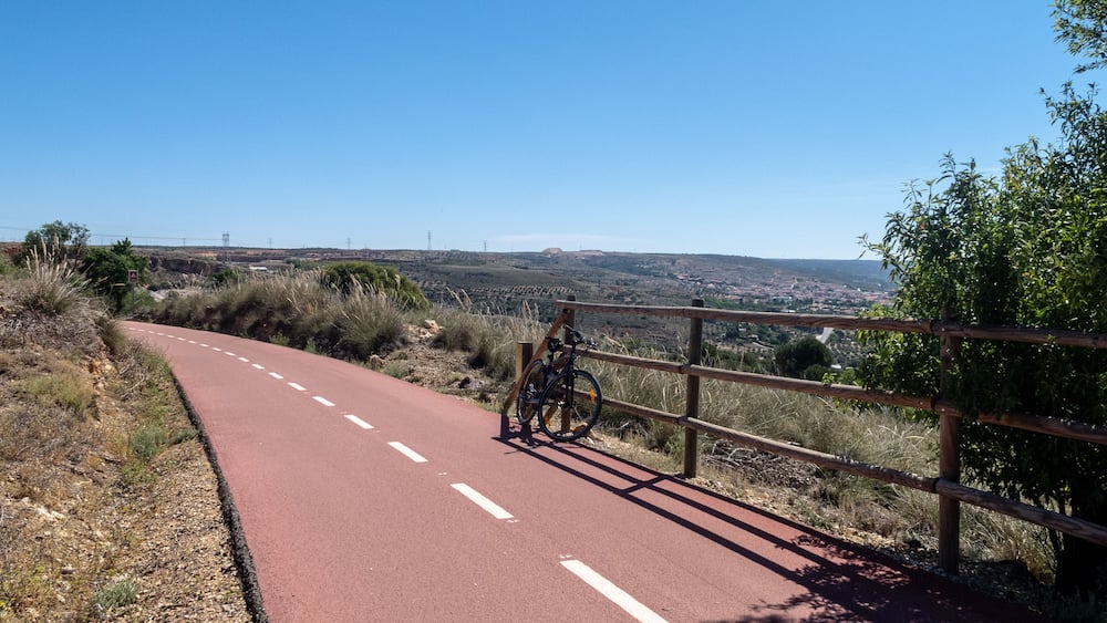 A paved road with a bicycle parked by a wooden fence, surrounded by dry vegetation and hills in the background.