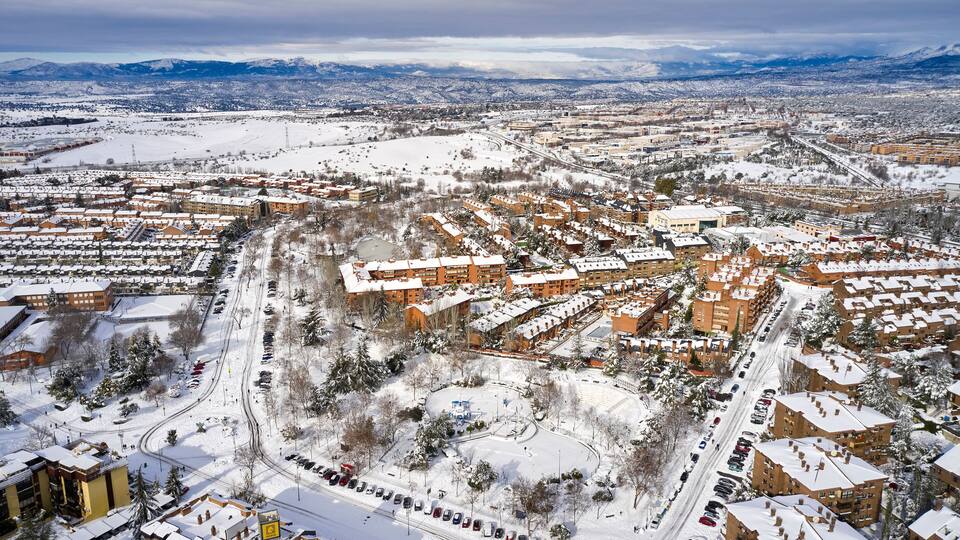 A snow-covered urban area with residential buildings, roads, and distant mountains.