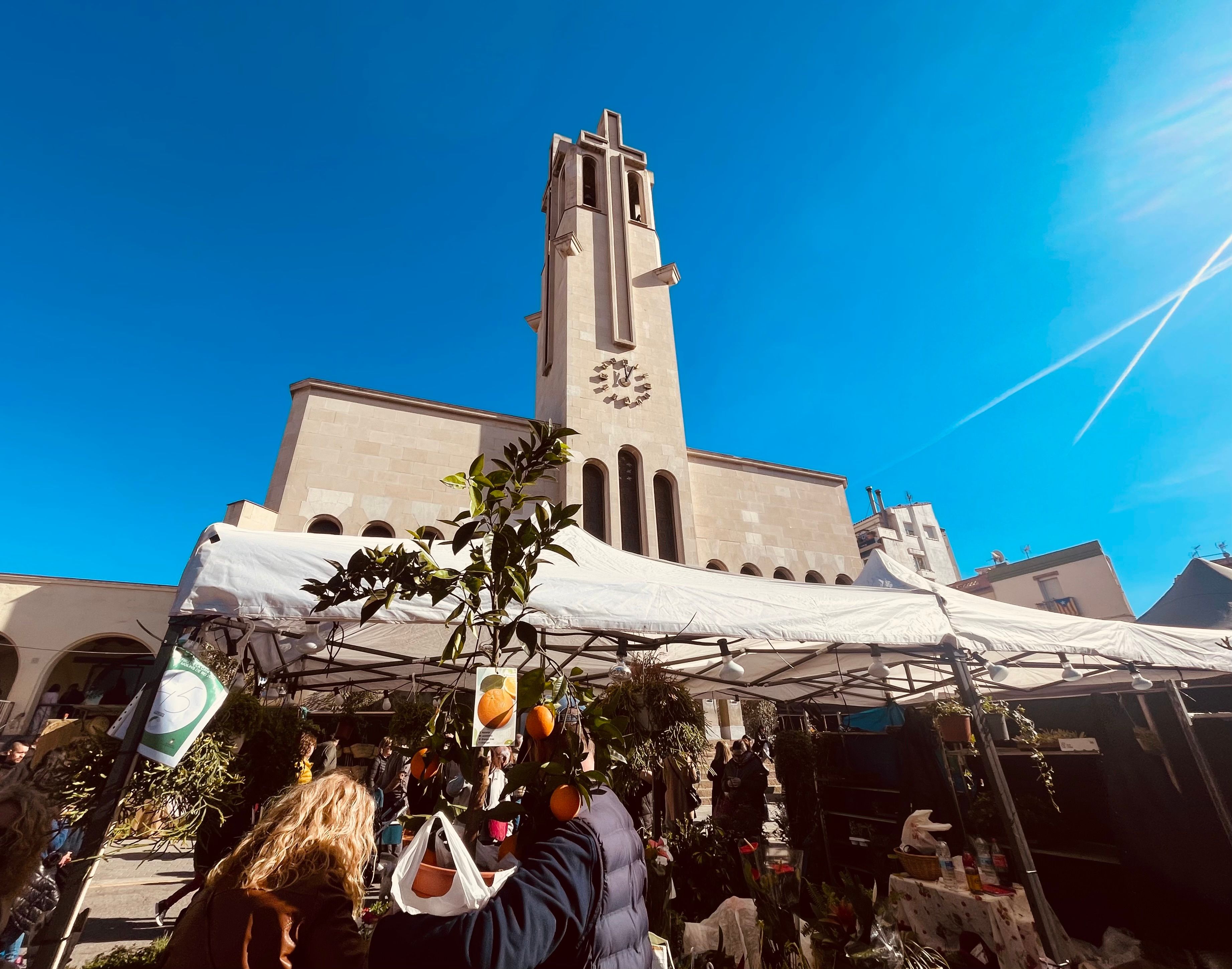 A church with a tall bell tower and a clock, surrounded by a market area with tents and people.