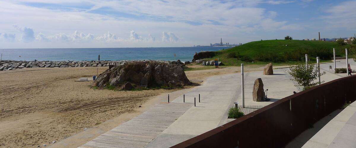 A beach with a wooden walkway, large rocks, and a grassy hill.