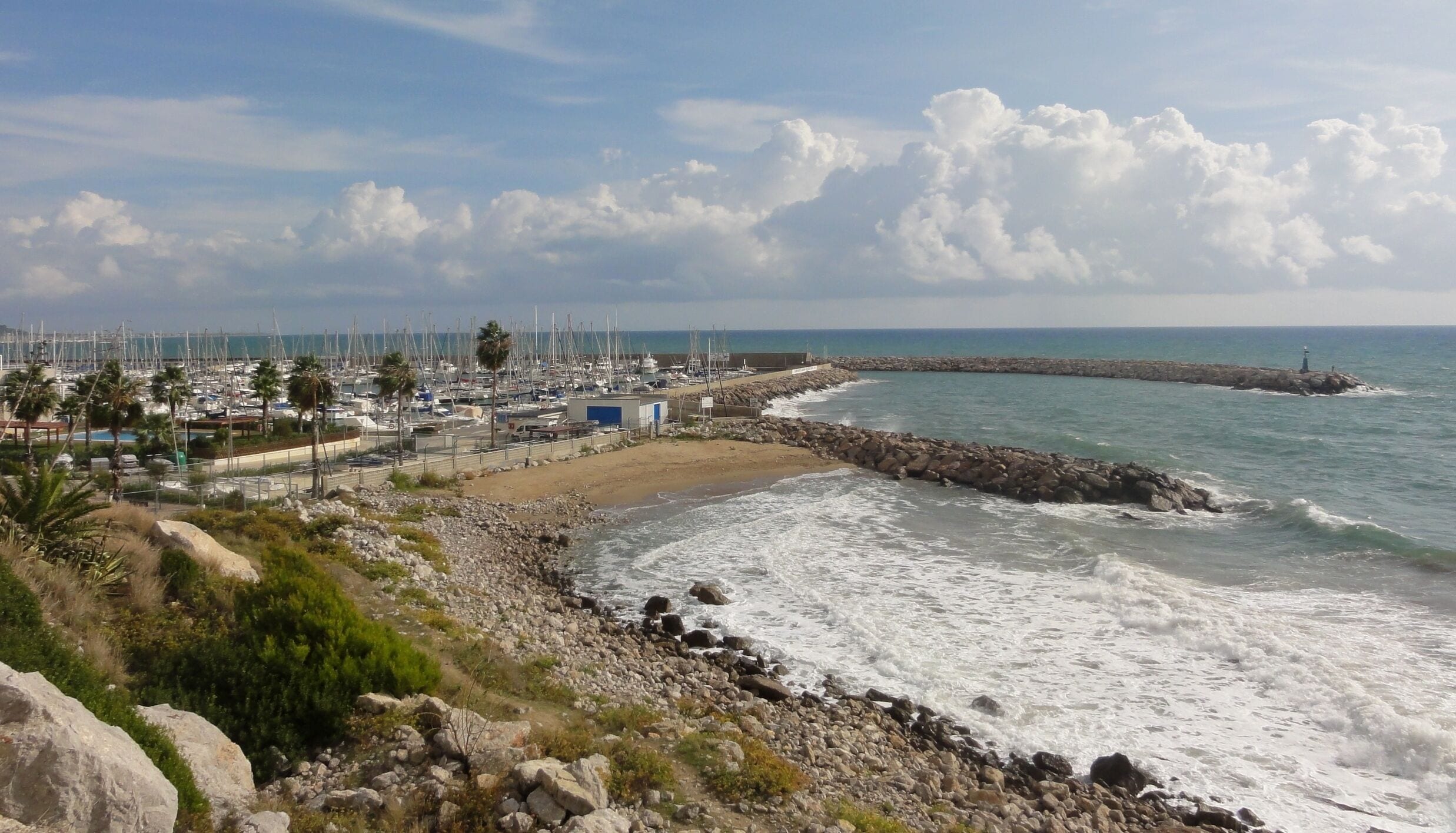 A coastal marina with boats, a rocky shoreline, and a lighthouse in the distance.
