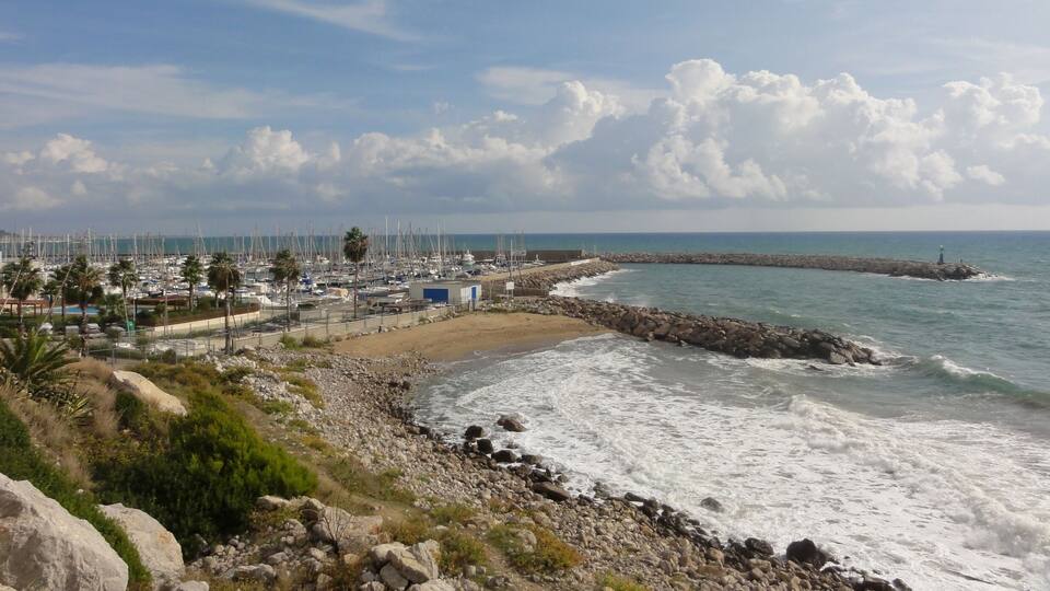 A coastal marina with boats, a rocky shoreline, and a lighthouse in the distance.
