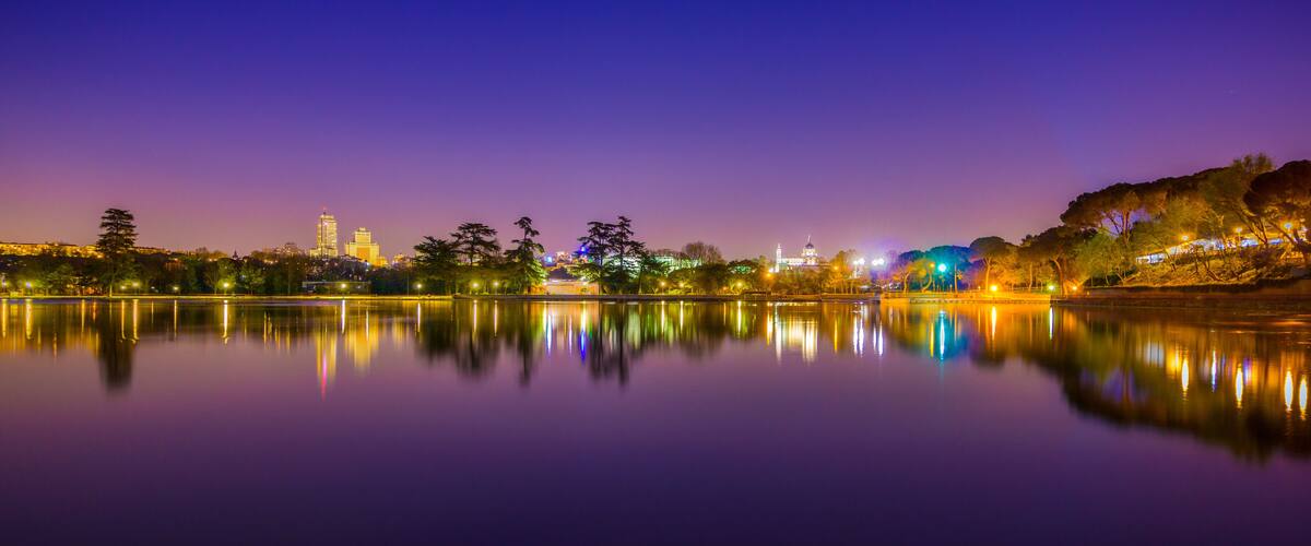 A calm lake reflecting city lights at dusk with a prominent building and trees along the shore.