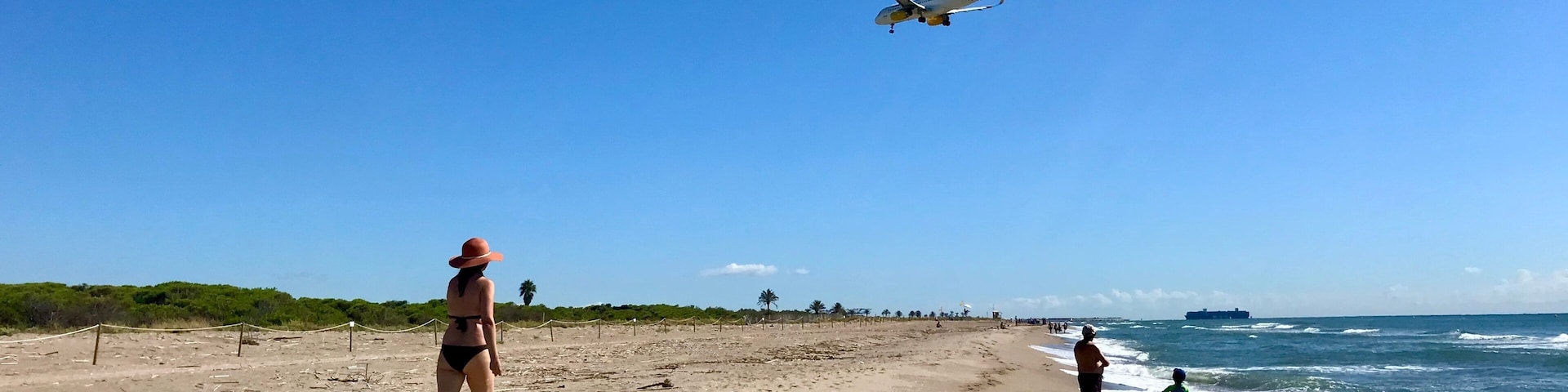 Una playa con un avión volando en lo alto, un barco en el agua y gente caminando por la orilla.