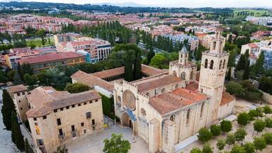 Una iglesia histórica con un campanario, rodeada por una plaza con árboles y vistas a un paisaje urbano de edificios con tejados rojos.