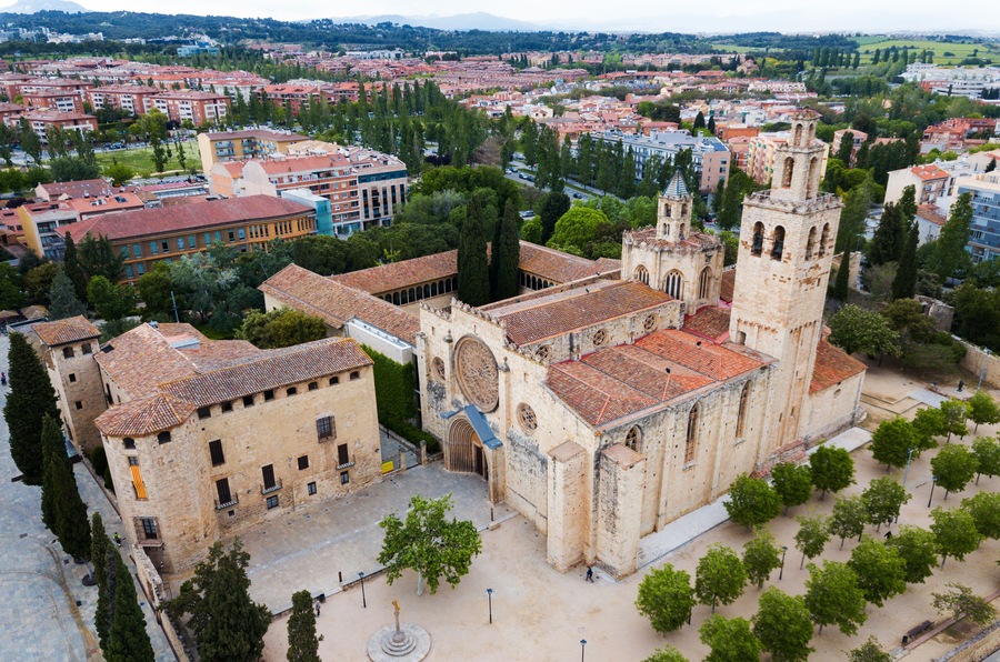 Una iglesia histórica con un campanario, rodeada por una plaza con árboles y vistas a un paisaje urbano de edificios con tejados rojos.