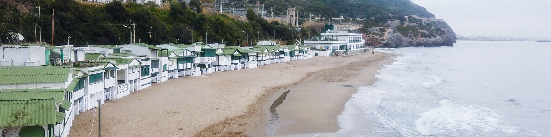 Uma praia com uma fileira de edifícios brancos com telhados verdes, um litoral rochoso e um fundo com colinas.