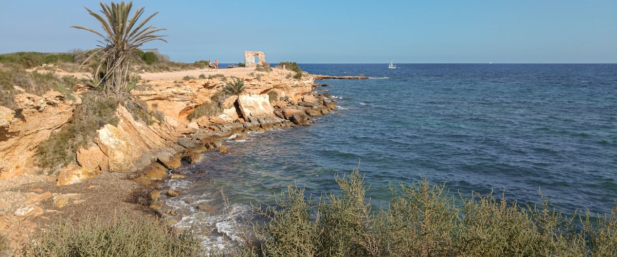 A rocky coastline with a palm tree, a small structure, and a sailboat in the distance.