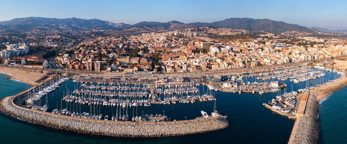 An aerial view of a coastal city with a marina filled with boats, a beach, and a mountainous backdrop.
