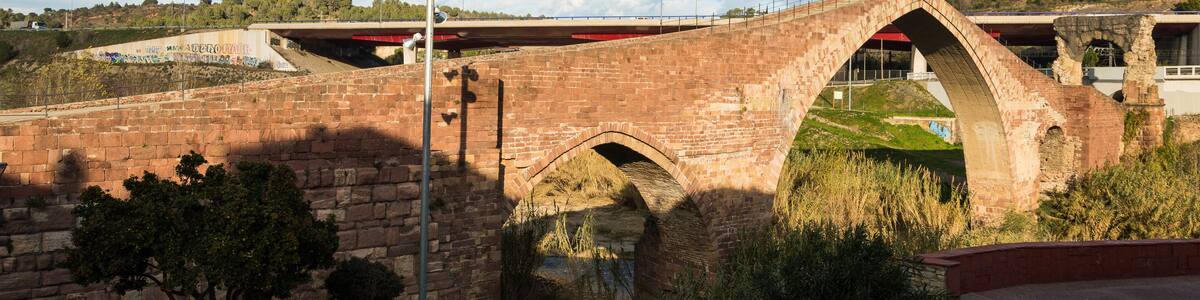 Un pont de pierre historique, orné d’arches, une zone herbeuse parsemée de rochers et un ciel dégagé.
