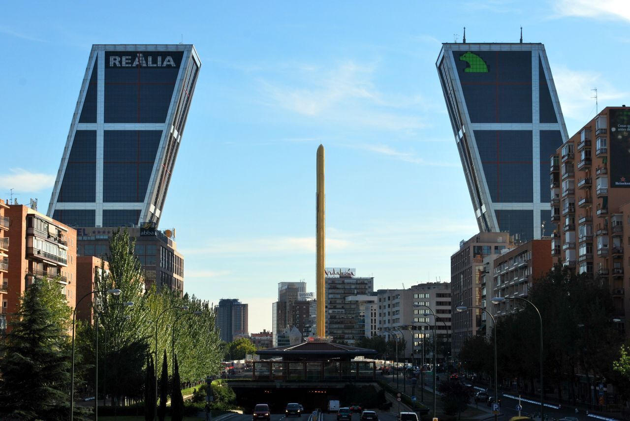 Two tall buildings with 'REALIA' signage, a tall monument, and a busy urban street with trees and parked cars.