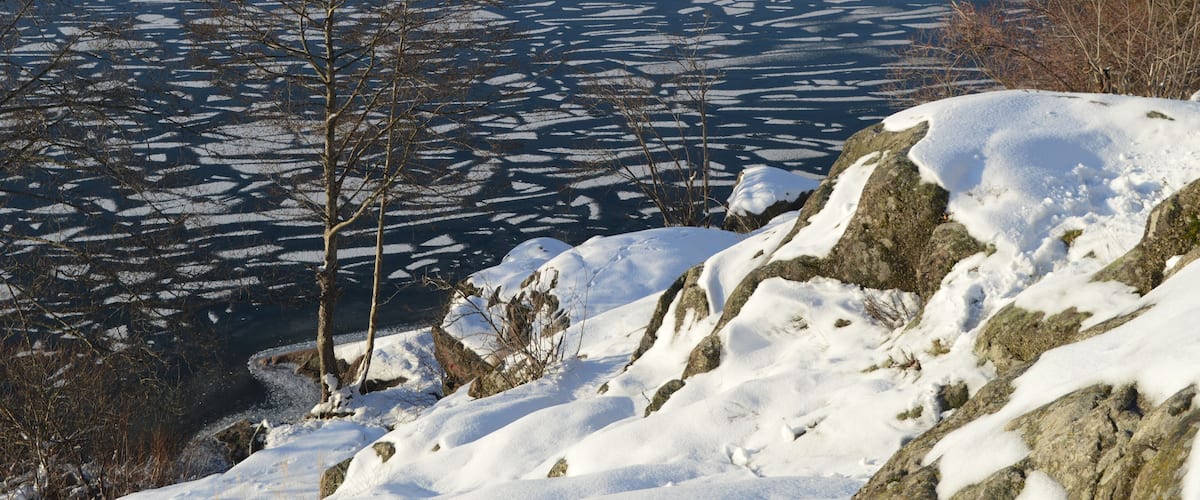A frozen lake with ice patterns, snow-covered rocks, and leafless trees.