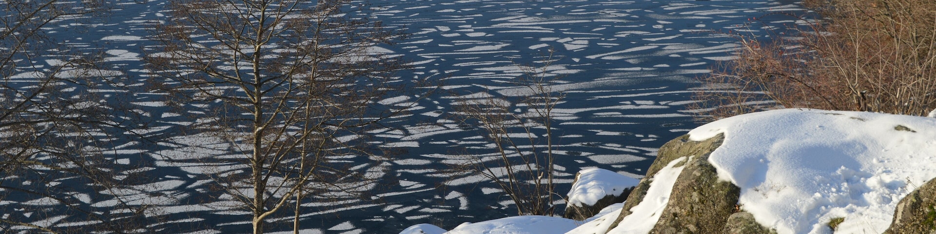 A frozen lake with ice patterns, snow-covered rocks, and leafless trees.
