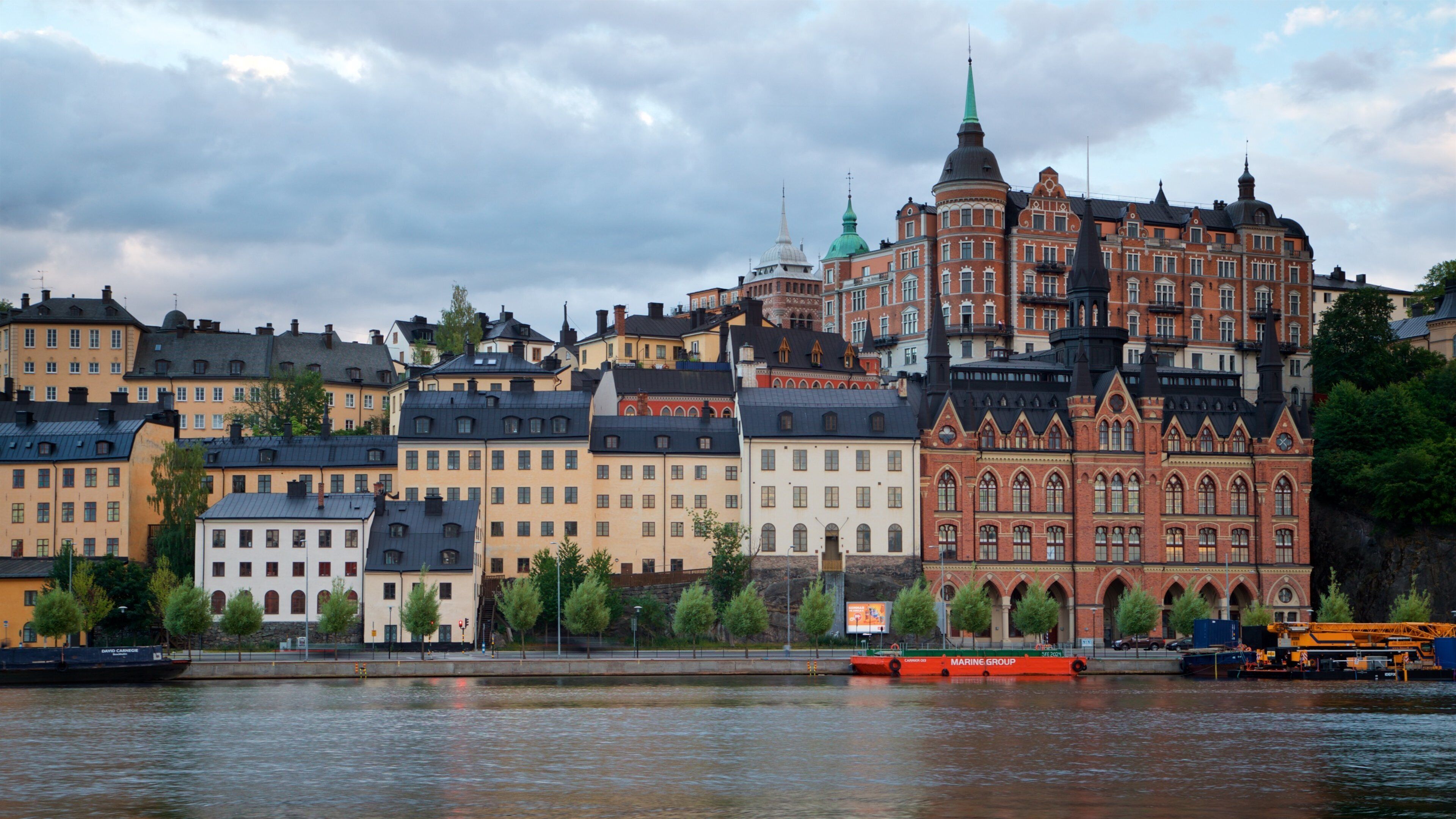A waterfront cityscape with historic buildings and a prominent red brick structure with a green spire.