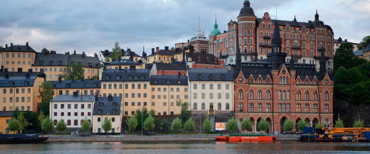 A waterfront cityscape with historic buildings and a prominent red brick structure with a green spire.