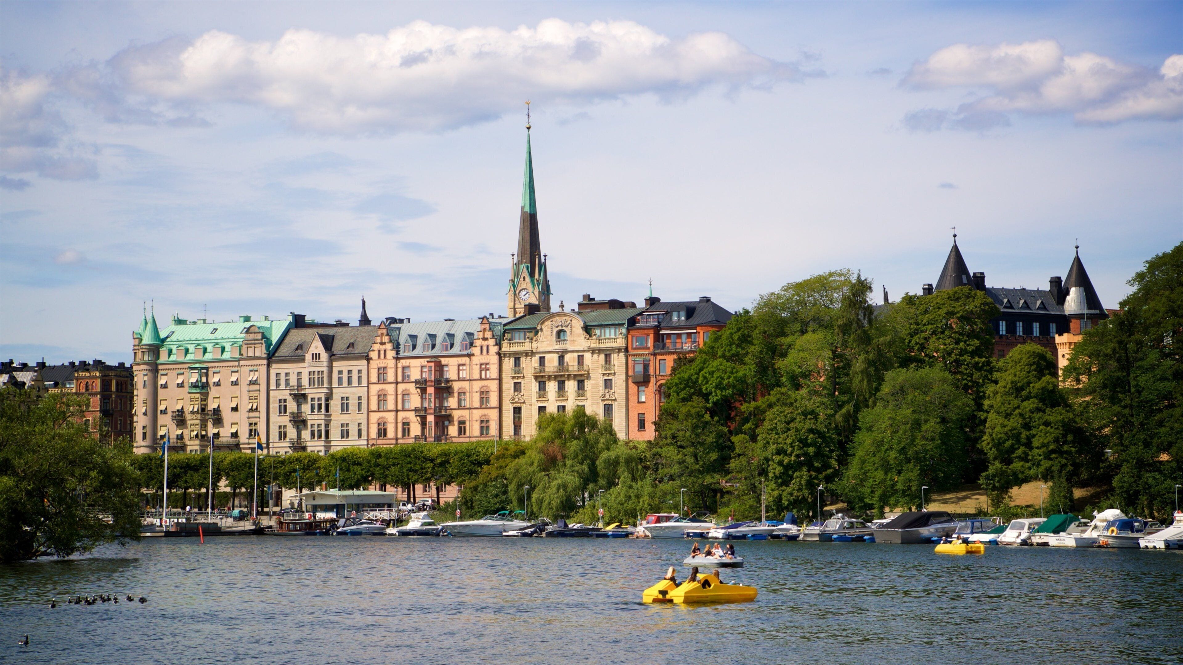 Ein Fluss mit Booten, darunter ein gelbes Boot im Vordergrund, und eine Stadtansicht mit historischen Gebäuden und einem Kirchturm im Hintergrund.