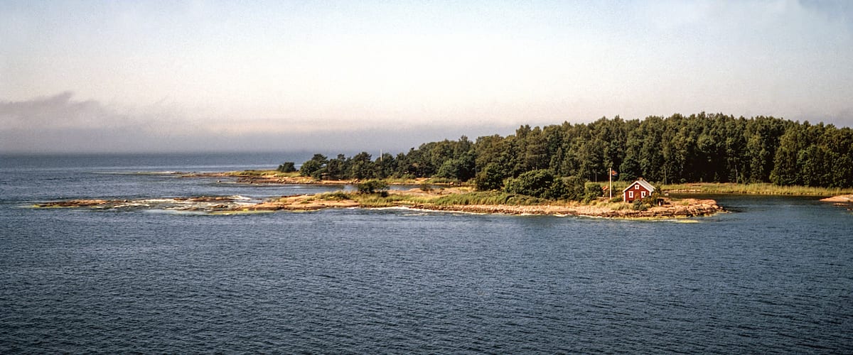 A coastal landscape with a small island featuring a red house surrounded by dense forest and calm waters.