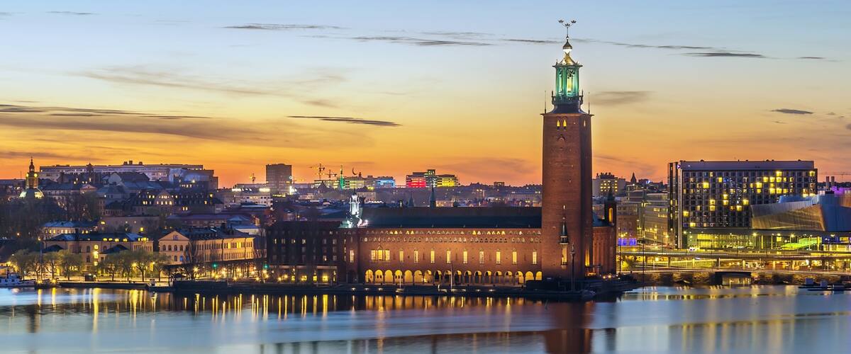 A cityscape at dusk with a prominent tower reflecting in the water, surrounded by illuminated buildings and a clear sky.