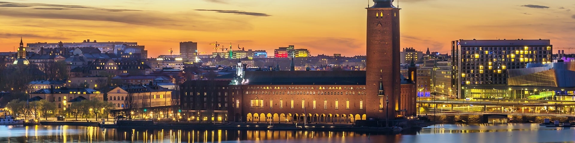 A cityscape at dusk with a prominent tower reflecting in the water, surrounded by illuminated buildings and a clear sky.