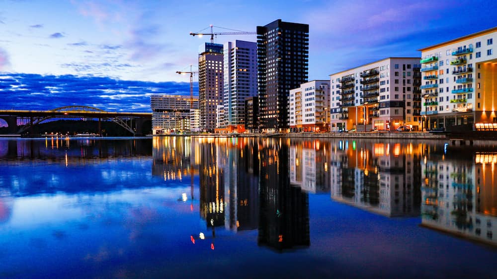 Eine Skyline mit modernen Wolkenkratzern, ein ruhiger Fluss, der die Gebäude spiegelt, und eine Brücke im Hintergrund.