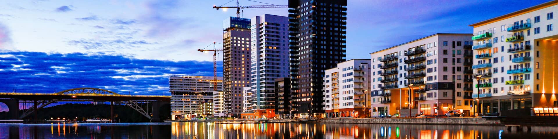 A cityscape with modern high-rise buildings, a calm river reflecting the structures, and a bridge in the background.