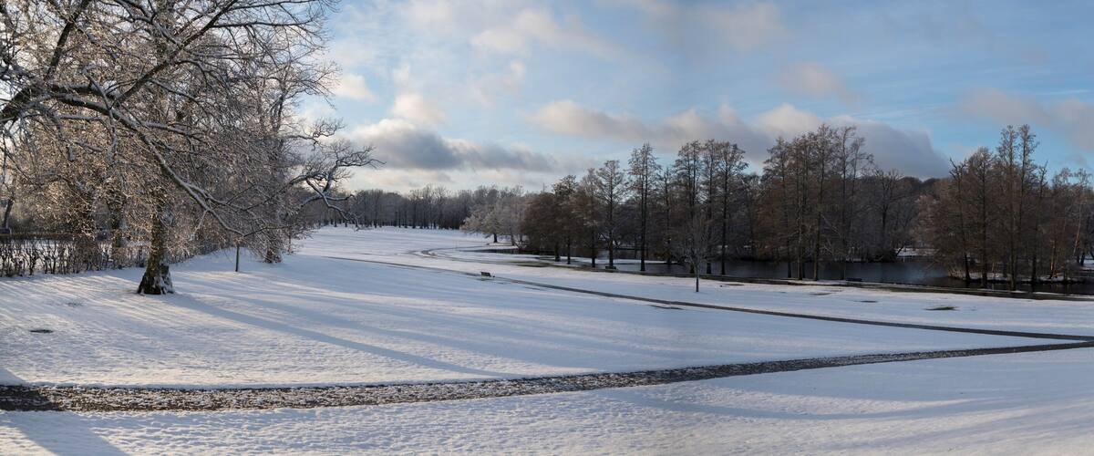 A snow-covered park with a river, trees, and a bench.