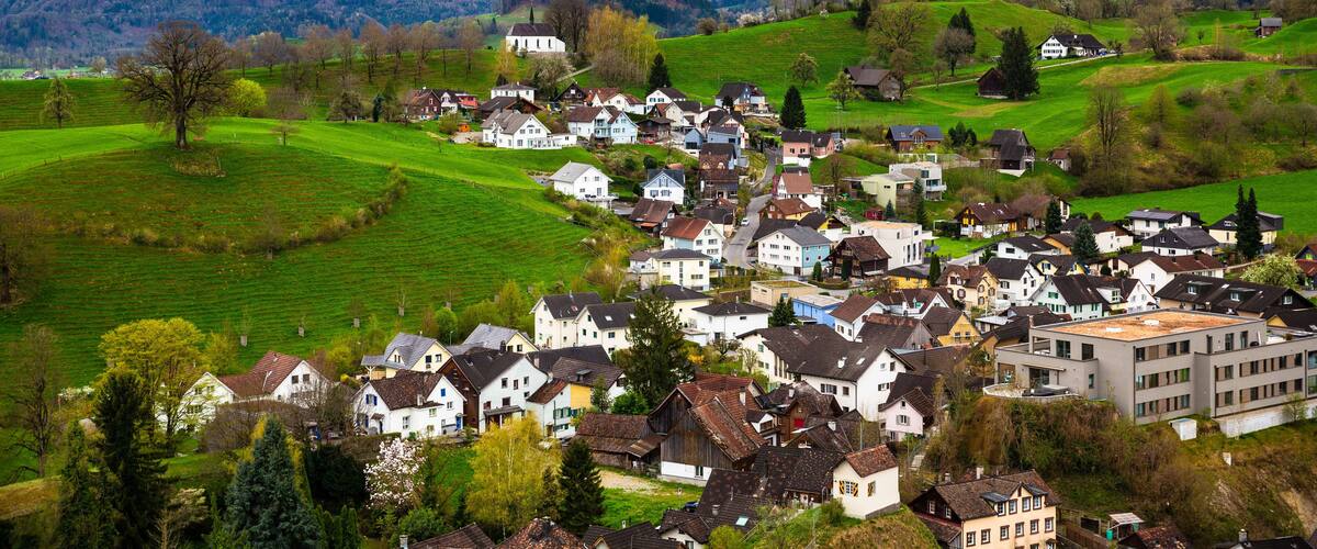 A village nestled in a valley with green fields and houses, surrounded by mountains.