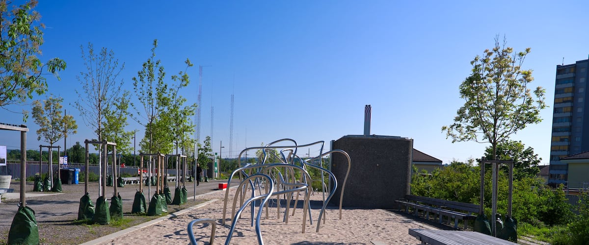 A sandy playground with metal exercise equipment, benches, and young trees.