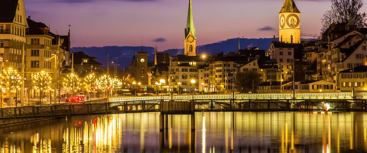 A cityscape at dusk with illuminated buildings, a bridge, and a river reflecting the lights.