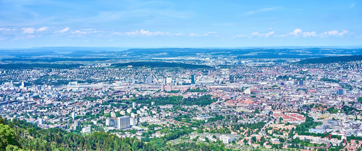 Laaja panoraamanäkymä kaupunkiin, jossa on tiheään rakennettuja asuinalueita, viheralueita ja kauempana kohoavia kukkuloita.