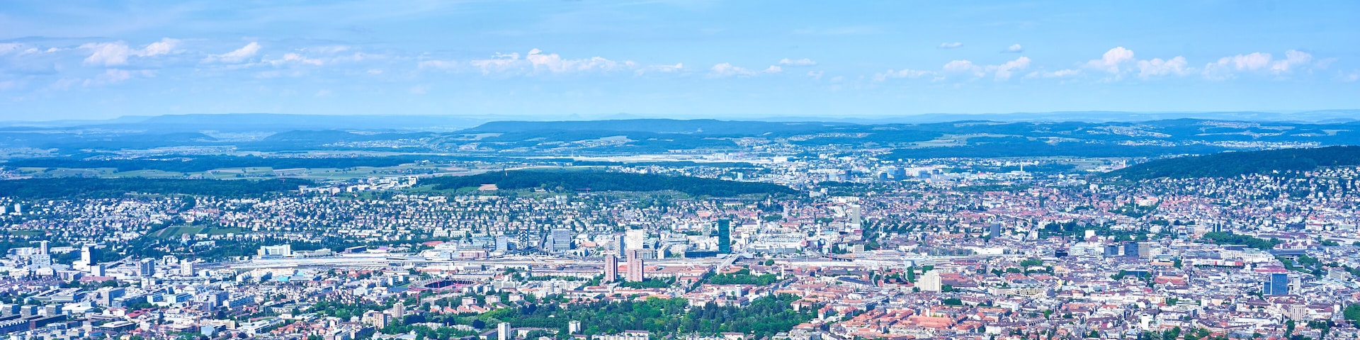 Vista panorámica de una ciudad con zonas urbanas densas, espacios verdes y colinas distantes.