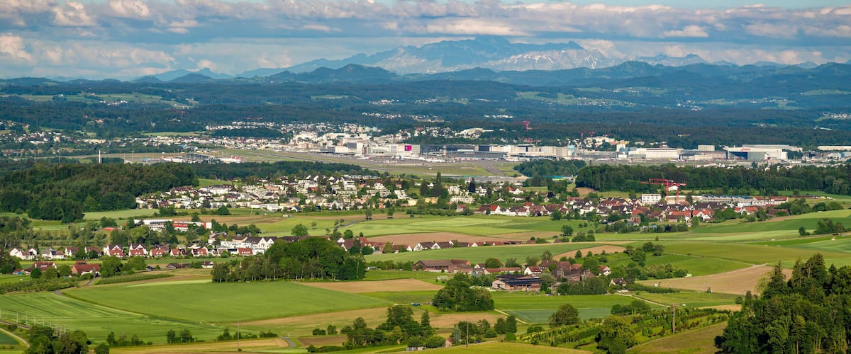 A town with a river, mountains in the background, and green fields.