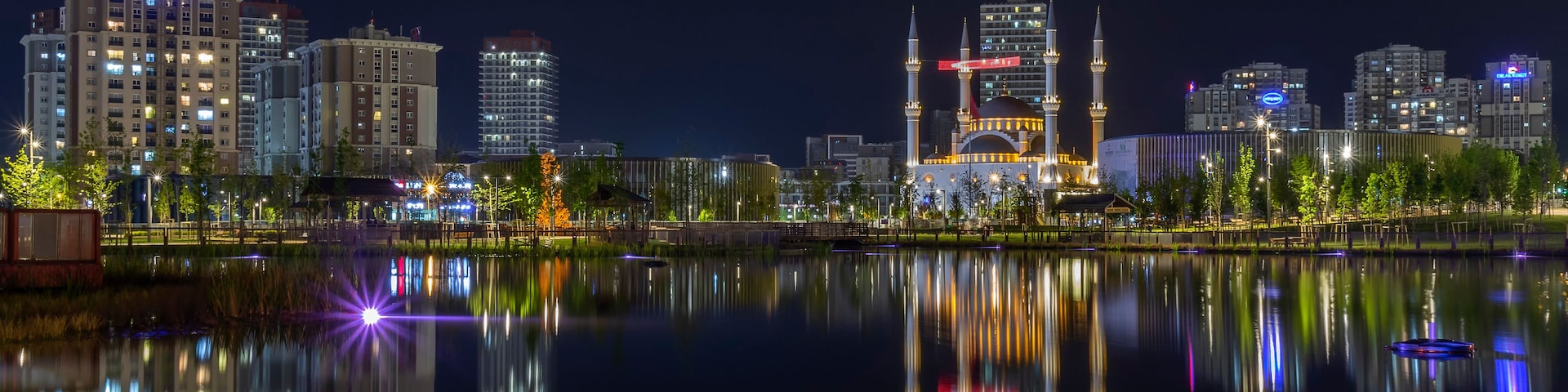 Paisagem urbana noturna com edifícios iluminados, uma mesquita com duas minaretes e um corpo d'água calmo refletindo as luzes.