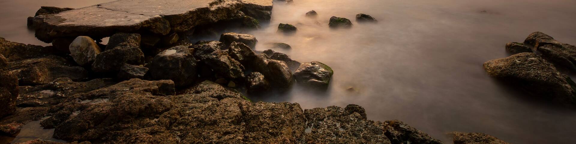 A calm body of water with a rocky shoreline, a wooden dock, and a distant coastline under a cloudy sky.
